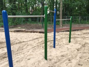 Drei farbige Reckstangen auf einem sandigen Spielplatz vor einem bewaldeten Hintergrund mit Gitterzaun.