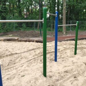 Drei farbige Reckstangen auf einem sandigen Spielplatz vor einem bewaldeten Hintergrund mit Gitterzaun.