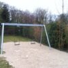 Zwei leere Schaukeln aus Metall auf einem Spielplatz mit Sand, umgeben von Bäumen und Sträuchern.