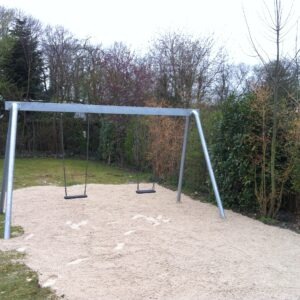 Zwei leere Schaukeln aus Metall auf einem Spielplatz mit Sand, umgeben von Bäumen und Sträuchern.