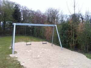 Zwei leere Schaukeln auf einem Spielplatz mit Sandboden, umgeben von Bäumen.