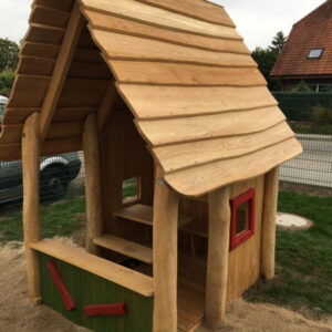 Holzspielhaus mit schrägem Dach, roten Fensterrahmen und grün-roter Wandverkleidung, auf Sand in einem Gartenbereich.