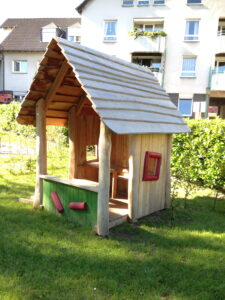 Holzspielhaus mit schrägem Dach, roten Fenstern und grüner Vorderseite vor einem Wohnhaus auf einer Wiese.