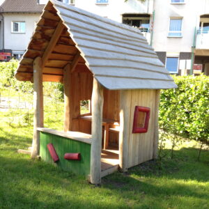 Holzspielhaus mit schrägem Dach, roten Fenstern und grüner Vorderseite vor einem Wohnhaus auf einer Wiese.