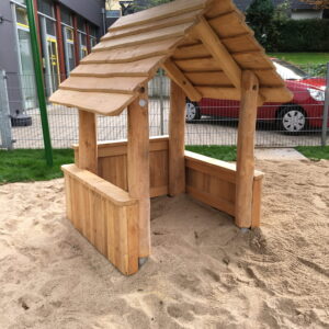 Holzhütte im Sandkasten eines Spielplatzes mit umliegenden Gebäuden und Bäumen im Hintergrund.