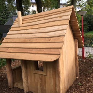 Holzhütte mit schrägem Dach und kleinem Fenster in einem Garten, umgeben von Bäumen und einem Zaun.