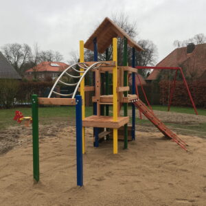 Bunter Spielplatz mit Klettergerüst, Rutsche und Schaukel auf Sand, umgeben von Wohnhäusern und Bäumen bei bewölktem Himmel.