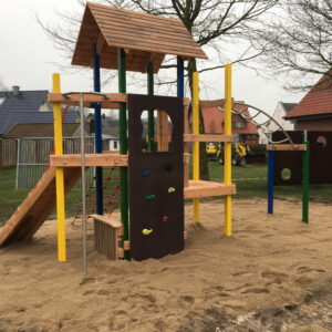 Holzspielplatz mit Sandboden, Klettergerüst, Kletterwand und Rutsche. Wohnhäuser und Bäume im Hintergrund.