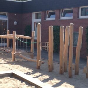 Holzklettergerüst auf einem Sandspielplatz vor einem Backsteingebäude mit mehreren Fenstern.