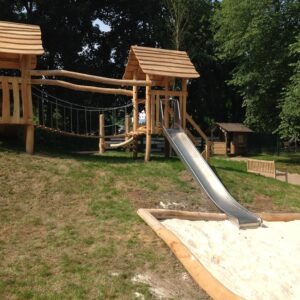 Holzspielplatz mit Rutsche, Hängebrücke und Sandkasten auf einem Hügel, umgeben von Bäumen und einer Bank im Hintergrund.