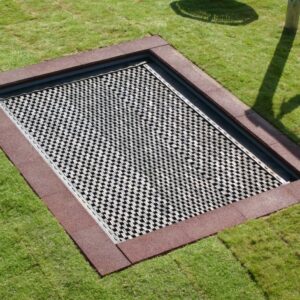 Bodentrampolin im Rasen, rechteckig, mit rotem Rand eingefasst. Ein Teil eines Schattens und Schuhe einer Person sind zu sehen.