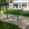 Grüne Klimmzugstangen auf einem sandigen Untergrund, umgeben von Gras, mit einem Gebäude im Hintergrund.