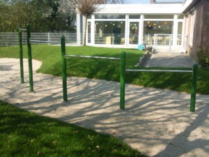 Grüne Klimmzugstangen auf einem sandigen Untergrund, umgeben von Gras, mit einem Gebäude im Hintergrund.