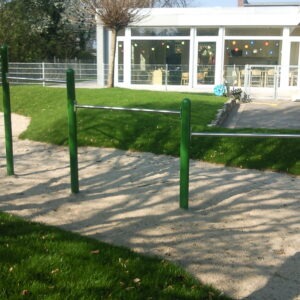 Grüne Klimmzugstangen auf einem sandigen Untergrund, umgeben von Gras, mit einem Gebäude im Hintergrund.