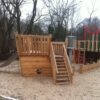 Holzspielplatz in Schiffsform mit Rutsche, Kletterseilen und Sandboden, umgeben von kahlen Bäumen und einem Gebäude im Hintergrund.
