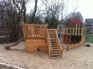 Holzspielplatz in Schiffsform mit Rutsche, Kletterseilen und Sandboden, umgeben von kahlen Bäumen und einem Gebäude im Hintergrund.