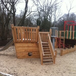 Holzspielplatz in Schiffsform mit Rutsche, Kletterseilen und Sandboden, umgeben von kahlen Bäumen und einem Gebäude im Hintergrund.